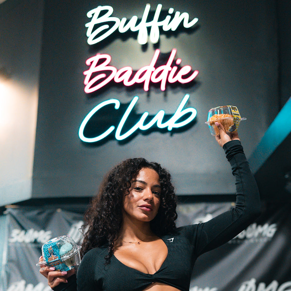A woman in a black crop top holds up two packaged muffins, standing in front of a neon sign that reads Buffin Baddie Club.