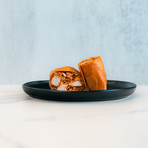 A black plate displays the Spicy Buffalo Wrap, cut in half to reveal its shredded meat filling. Set on a white marble surface with a light gray background, this scene is perfect inspiration for healthy wraps and meal prep ideas.