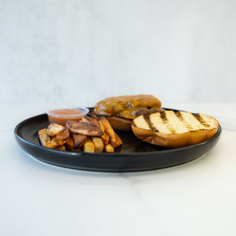 The Stacked and Jacked features a grilled hamburger bun, high-protein grass-fed beef cheeseburger patty, thick Idaho fries, and a dipping sauce, all served on a black plate atop a white surface.