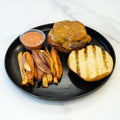 A black plate holds the Stacked and Jacked high-protein grass-fed beef cheeseburger with the top bun on the side, served with crispy Idaho fries and dipping sauce, all set on a white marble surface.
