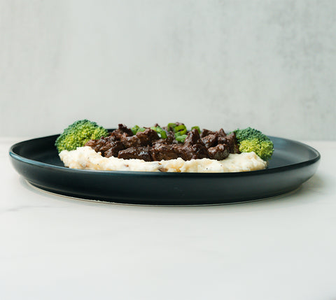 The Herb Butter Steak Tips Bowl features tender steak tips over mashed potatoes with a savory herb butter sauce, garnished with green onions and served alongside steamed broccoli—a tasty, high-protein meal prep option.