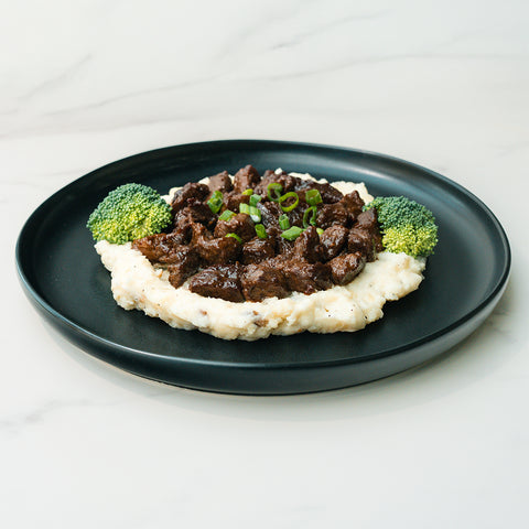 The Herb Butter Steak Tips Bowl features mashed potatoes topped with steak beef stew and green onions, plus broccoli on the side—a delicious, high-protein meal prep option served on a black plate.