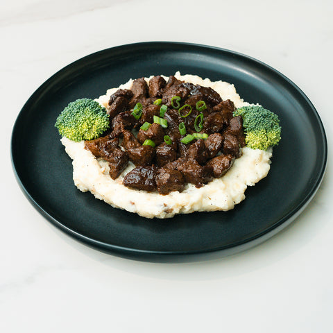 The Herb Butter Steak Tips Bowl features mashed potatoes topped with grass-fed steak in gravy, garnished with green onions and broccoli florets—a hearty, high-protein meal prep option.