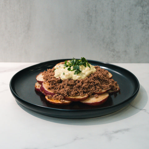 The Street Corn Beef Bowl features ground beef, sliced potatoes, creamy sauce, and herbs for a high-protein meal, served on a black plate atop a white marble surface with a light gray background.