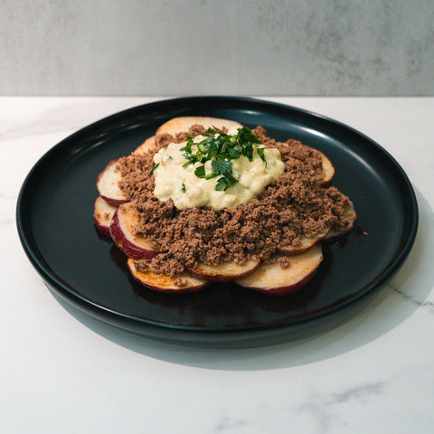 The Street Corn Beef Bowl features thinly sliced red potatoes, ground beef, creamy sauce, and fresh herbs for a macro-friendly, high-protein meal prep. Presented on a black plate atop a white marble surface.