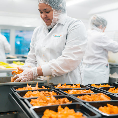 A worker in a white coat, hairnet, and gloves portions orange food into black trays in a food processing facility. Another worker is blurred in the background.