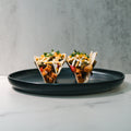 Two Mexican Tacos filled with diced vegetables and high-protein chicken, topped with avocado ranch and chopped herbs, are served on a black plate against a light gray background.