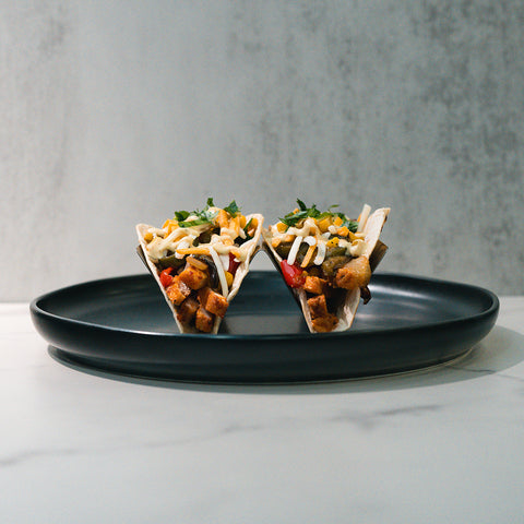Two Mexican Tacos filled with diced vegetables and high-protein chicken, topped with avocado ranch and chopped herbs, are served on a black plate against a light gray background.