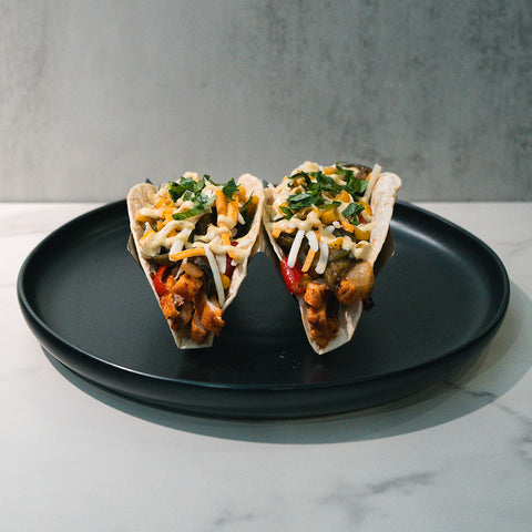 The Mexican Taco features two soft tacos filled with grilled chicken, sautéed vegetables, shredded cheese, and fresh herbs, served on a black plate against a neutral background.