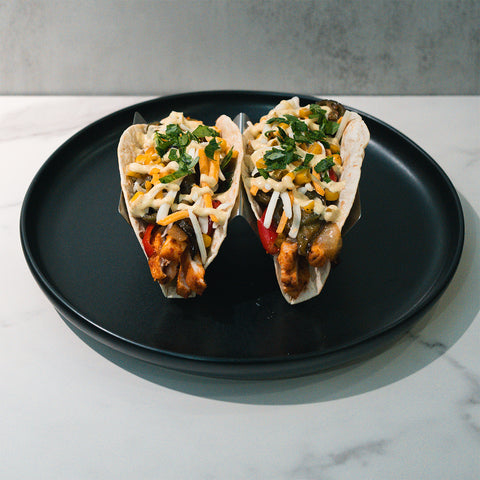 The Mexican Taco features two soft tacos filled with grilled chicken, sautéed vegetables, shredded cheese, and fresh herbs, served upright on a black plate over a light marble background for a delicious high-protein meal.