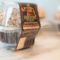 A close-up of a Buffin Muffin - German Chocolate Cake in a plastic container shows its shredded coconut topping. The high-protein muffin is a fitness-friendly treat, with nutritional info and branding clearly visible on the label.