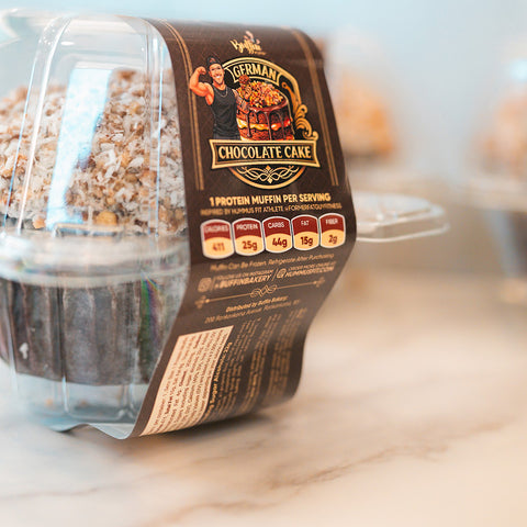A close-up of a Buffin Muffin - German Chocolate Cake in a plastic container shows its shredded coconut topping. The high-protein muffin is a fitness-friendly treat, with nutritional info and branding clearly visible on the label.