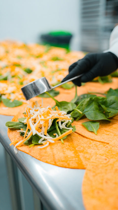 A gloved hand uses a metal scoop to add shredded cheese to spinach on orange tortillas, lined up on a metal counter in a food preparation setting.