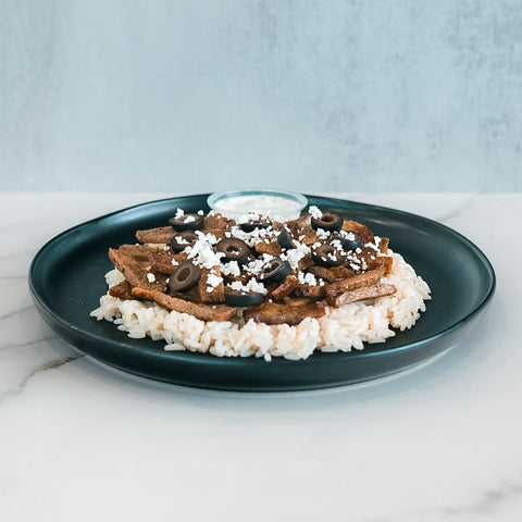 An Olympian Chicken Gyro Bowl featuring a black plate with rice, sliced meat, black olives, and crumbled white cheese, served with a side of white sauce.
