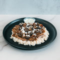 An Olympian Chicken Gyro Bowl with sliced meat, crumbled white cheese, and sliced black olives served over rice on a black plate with a side of white sauce.