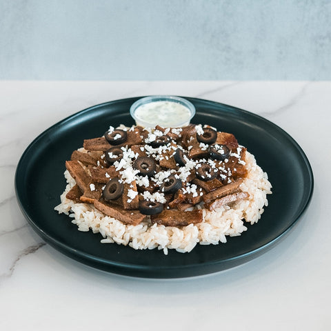 An Olympian Chicken Gyro Bowl with sliced meat, crumbled white cheese, and sliced black olives served over rice on a black plate with a side of white sauce.