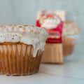 A close-up of the VIP Buffin Muffin - Cookie Butter Tres Leches, topped with icing and infused with cookie butter, sits on a white surface. A packaged muffin appears blurred in the background.