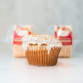A single VIP Buffin Muffin - Cookie Butter Tres Leches with white icing and cinnamon sits on a white surface, made with low-fat yogurt. Two packaged muffins are blurred in the background.