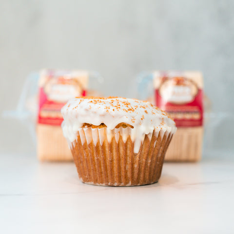 A single VIP Buffin Muffin - Cookie Butter Tres Leches with white icing and cinnamon sits on a white surface, made with low-fat yogurt. Two packaged muffins are blurred in the background.