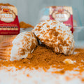 Two VIP Buffin Muffin - Cookie Butter Tres Leches pastries, coated in white icing and brown crumbs, rest on a crumb-covered surface, with blurred red-labeled packaging in the background.