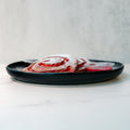 A black plate holding two red and white swirled pastries dusted with powdered sugar, with two small containers of red dipping sauce, on a white surface with a light grey background.
