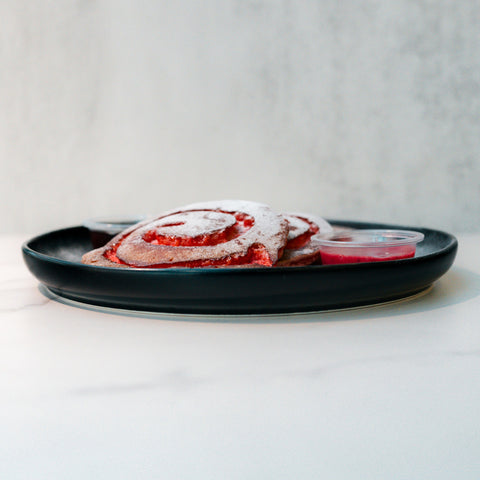 A black plate holding two red and white swirled pastries dusted with powdered sugar, with two small containers of red dipping sauce, on a white surface with a light grey background.