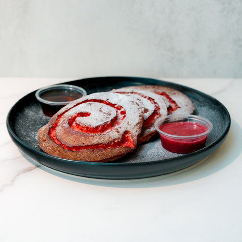 A black plate holds two large, swirled pastries dusted with powdered sugar, accompanied by two small containers of red and brown dipping sauces, on a white marble surface.