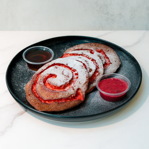 Three cinnamon rolls with red filling and powdered sugar are served on a black plate with two small cups of dipping sauces, one dark and one red, on a white marble surface.