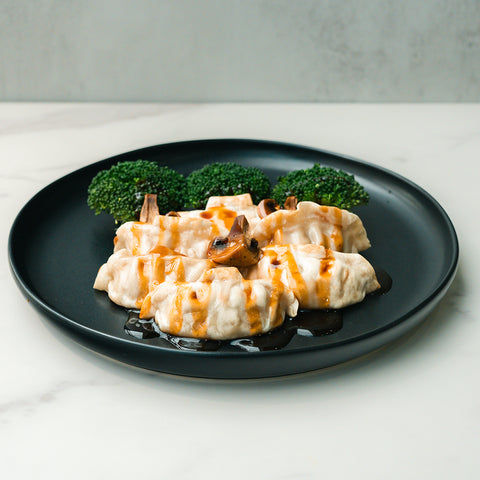 Five Chicken Mushroom Pot Stickers drizzled with teriyaki sauce are arranged on a black plate, with mushrooms and broccoli florets in the background, set on a white marble surface.