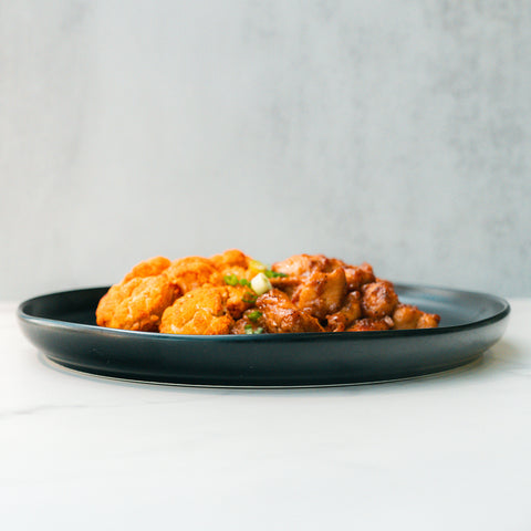 Bang Bang Chicken served on a black plate with crispy fritters and chopped green onion is a high-protein meal, ideal for pairing with Thai chili sauce, presented against a light, neutral background.