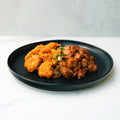 Bang Bang Chicken: A black plate with golden crispy fried chicken and saucy, brown Bang Bang Chicken garnished with green onions and Thai chili sauce, served on a white surface.