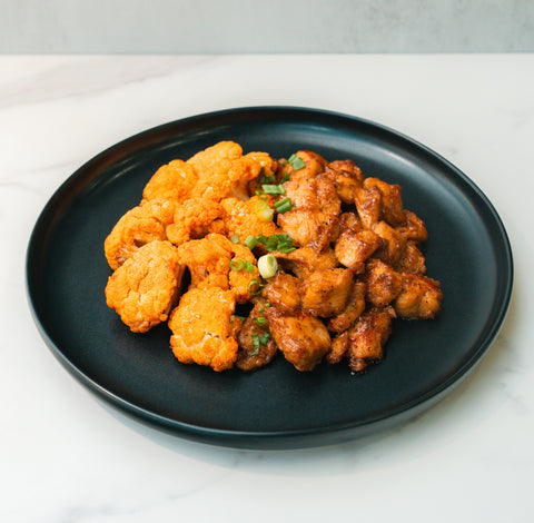 Bang Bang Chicken: Seasoned cauliflower and chicken chunks tossed in Thai chili sauce, garnished with green onions, served on a black plate atop white marble.
