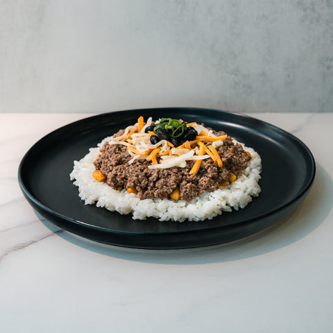 The Honey Garlic Beef Bowl features seasoned ground beef, white rice, cheese, black olives, and green onions for a colorful high-protein meal prep served in a black plate—perfect for your next healthy beef bowl on a marble surface.