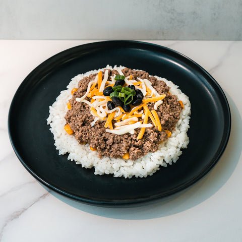 The Honey Garlic Beef Bowl features white rice, seasoned ground beef, shredded cheese, green onions, and black beans—a high-protein meal prep option served on a black plate over a white marble surface.