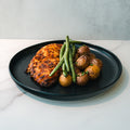 The Baked Herb Chicken and Potatoes meal features chicken breast, roasted baby potatoes, and green beans on a black plate, offering a high-protein meal prep option on a white marble surface.