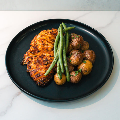Baked Herb Chicken and Potatoes—a high-protein meal prep with roasted baby potatoes and green beans, served on a black plate atop white marble.