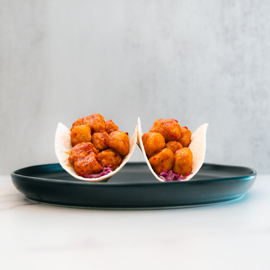 Two small tacos filled with honey garlic crispy chicken and purple cabbage are arranged upright on a dark plate against a light background, offering a flavorful twist perfect for high-protein meal prep enthusiasts.