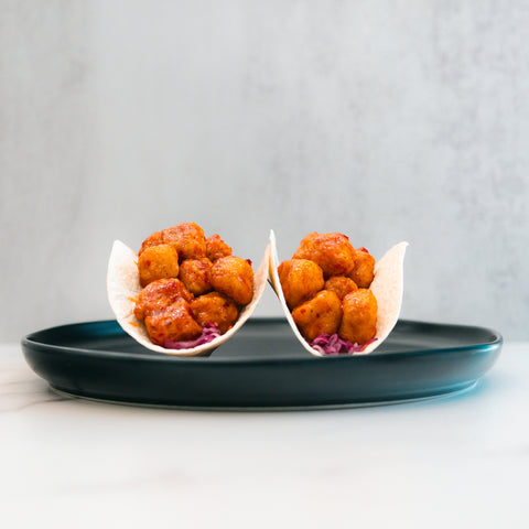 Two small tacos filled with honey garlic crispy chicken and purple cabbage are arranged upright on a dark plate against a light background, offering a flavorful twist perfect for high-protein meal prep enthusiasts.