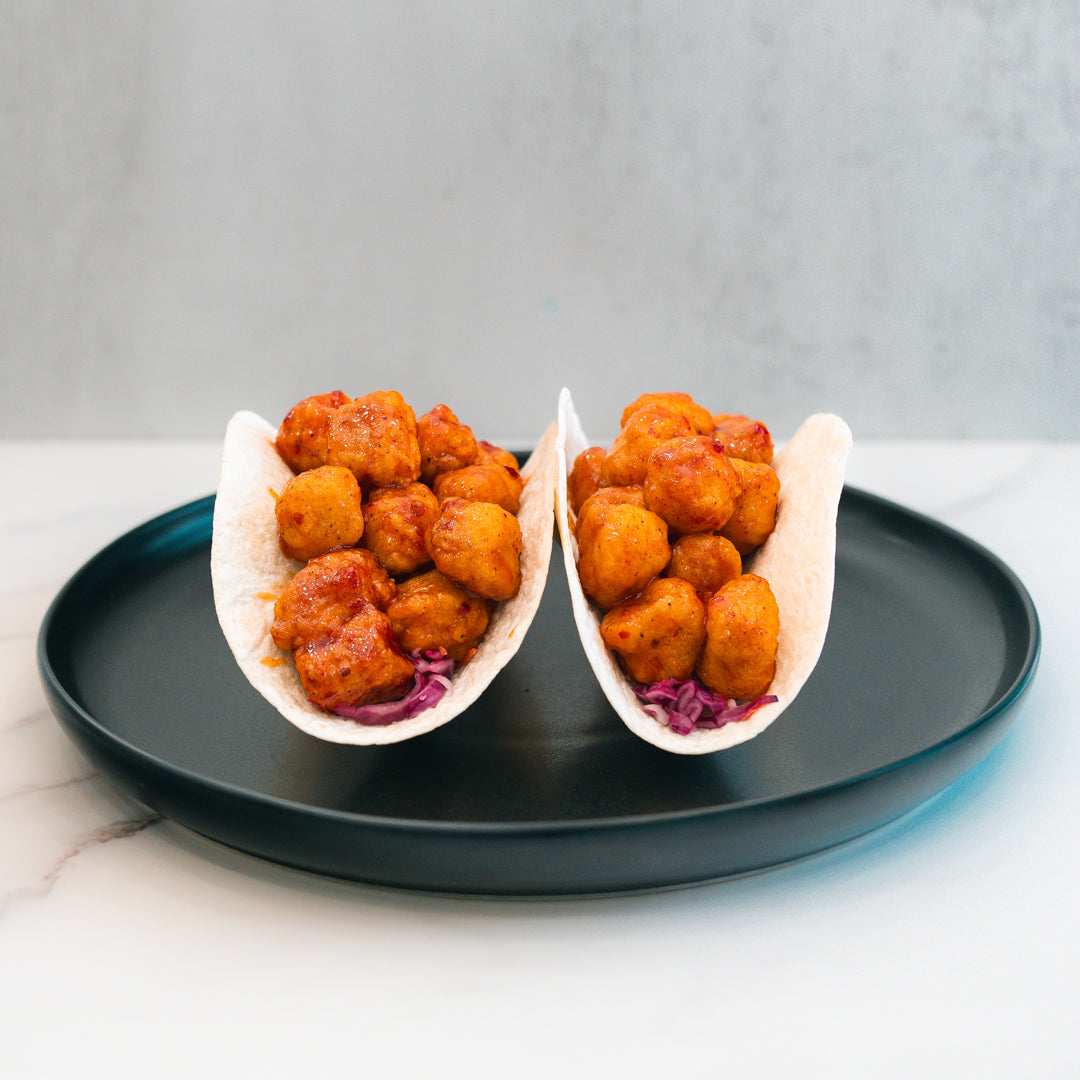 Enjoy the Honey Garlic Crispy Chicken Tacos—two soft taco shells filled with seasoned, golden-brown crispy chicken and shredded purple cabbage, served on a black plate against a light background. This irresistible taco delivers the perfect fusion of honey garlic flavor and classic crunch, making it an ideal high-protein meal option for your next food prep session.