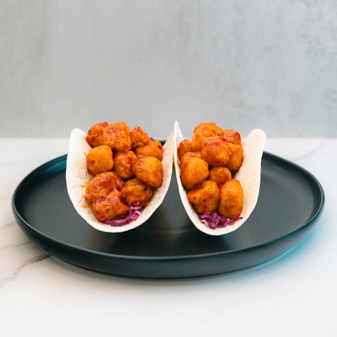 Enjoy the Honey Garlic Crispy Chicken Tacos—two soft taco shells filled with seasoned, golden-brown crispy chicken and shredded purple cabbage, served on a black plate against a light background. This irresistible taco delivers the perfect fusion of honey garlic flavor and classic crunch, making it an ideal high-protein meal option for your next food prep session.
