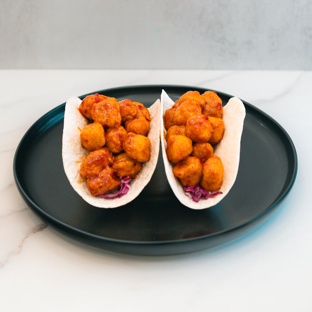 Two soft tacos filled with crispy, golden-brown pieces of honey garlic chicken and shredded purple cabbage sit on a black plate atop a white surface, making these Honey Garlic Crispy Chicken Tacos a delicious choice for high-protein meal prep.