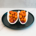 Two soft tacos filled with crispy, golden-brown pieces of honey garlic chicken and shredded purple cabbage sit on a black plate atop a white surface, making these Honey Garlic Crispy Chicken Tacos a delicious choice for high-protein meal prep.