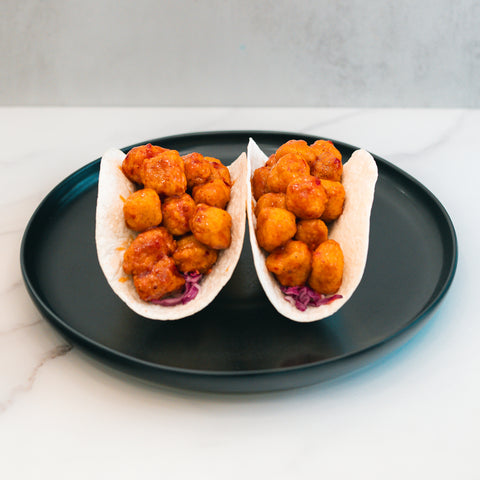 Two soft tacos filled with crispy, golden-brown pieces of honey garlic chicken and shredded purple cabbage sit on a black plate atop a white surface, making these Honey Garlic Crispy Chicken Tacos a delicious choice for high-protein meal prep.