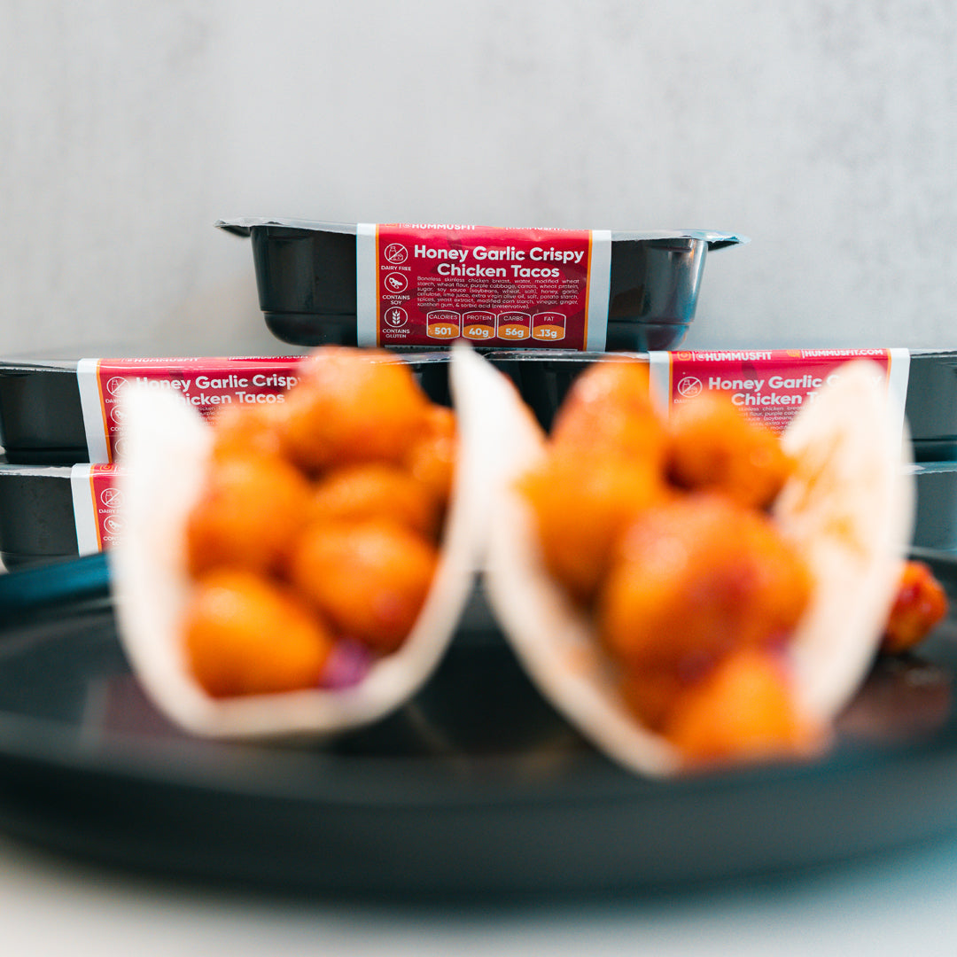 Two soft tacos filled with crispy chicken pieces are served on a black plate, showcasing the tempting Honey Garlic Crispy Chicken Tacos. In the background, black meal containers labeled “Honey Garlic Crispy Chicken Tacos” emphasize this High-Protein Meal Prep favorite. The brightly lit image keeps the focus softly on the delicious tacos.