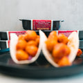 Two soft tacos filled with crispy chicken pieces are served on a black plate, showcasing the tempting Honey Garlic Crispy Chicken Tacos. In the background, black meal containers labeled “Honey Garlic Crispy Chicken Tacos” emphasize this High-Protein Meal Prep favorite. The brightly lit image keeps the focus softly on the delicious tacos.