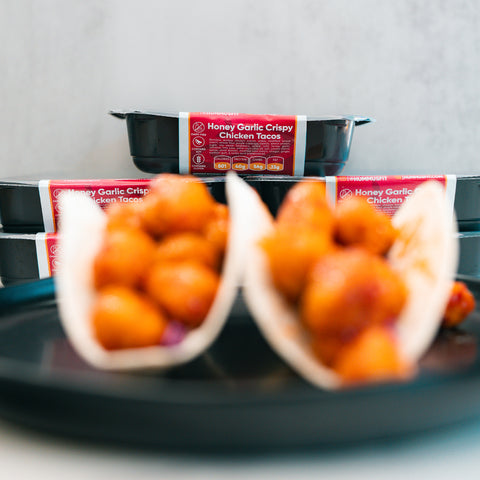 Two soft tacos filled with crispy chicken pieces are served on a black plate, showcasing the tempting Honey Garlic Crispy Chicken Tacos. In the background, black meal containers labeled “Honey Garlic Crispy Chicken Tacos” emphasize this High-Protein Meal Prep favorite. The brightly lit image keeps the focus softly on the delicious tacos.