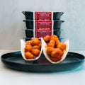 Two tacos filled with Honey Garlic Crispy Chicken rest on a black plate, while four stacked meal containers labeled Honey Garlic Crispy Chicken Tacos sit in the background—an ideal choice for high-protein meal prep.