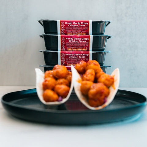 Two tacos filled with Honey Garlic Crispy Chicken rest on a black plate, while four stacked meal containers labeled Honey Garlic Crispy Chicken Tacos sit in the background—an ideal choice for high-protein meal prep.
