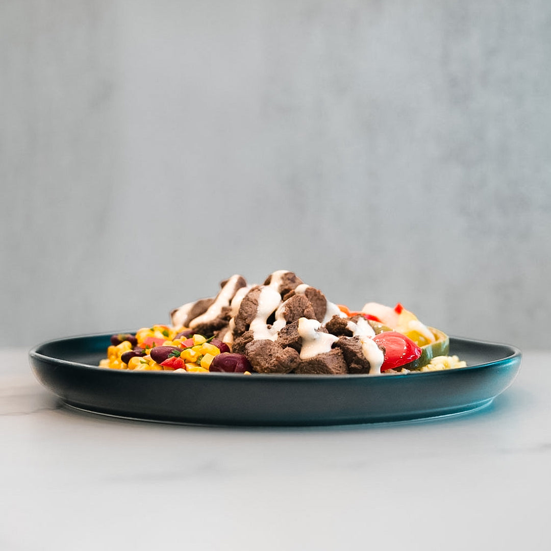 A dark plate displays a Texas Queso Steak Bowl—grilled steak topped with creamy white queso—accompanied by a vibrant combination of corn, beans, and cherry tomatoes. Set on a light surface with a neutral background, this high-protein meal is satisfying and naturally gluten-free.