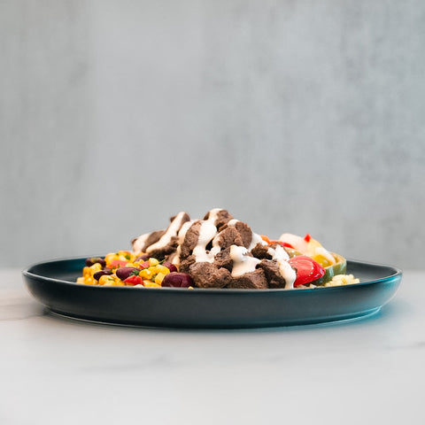 A dark plate displays a Texas Queso Steak Bowl—grilled steak topped with creamy white queso—accompanied by a vibrant combination of corn, beans, and cherry tomatoes. Set on a light surface with a neutral background, this high-protein meal is satisfying and naturally gluten-free.